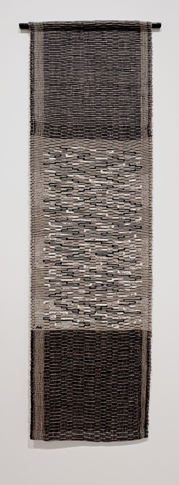 A large textile wall hanging made from white, black and silver threads. Threads weave in and out creating a random pattern of meandering threads.